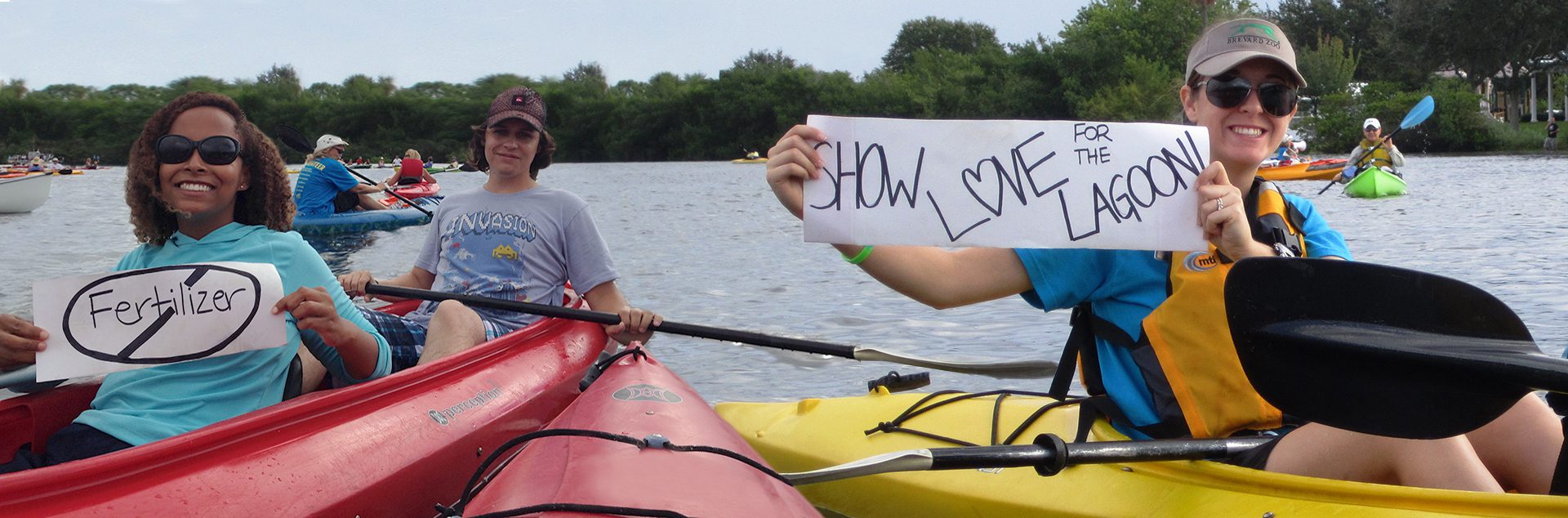 Kayakers, one of them holding up a "Show Love to the Lagoon" sign. Another holding a "No fertilizer" sign. They're smiling, assuming they're happy.